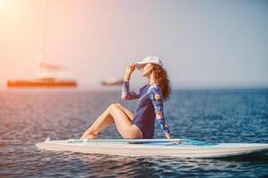 Woman sup yoga. Middle age sporty woman practising yoga pilates on paddle sup surfboard. Female stretching doing workout on sea water. Modern individual female hipster outdoor summer sport activity. photo