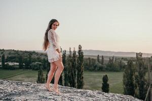 Happy woman in white boho dress on sunset in mountains. Romantic woman with long hair standing with her back on the sunset in nature in summer with open hands. Silhouette. Nature. Sunset. photo