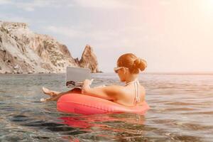 mujer persona de libre dedicación trabajos en ordenador portátil nadando en mar en rosado inflable anillo. contento turista en Gafas de sol flotante en inflable rosquilla y trabajando en ordenador portátil computadora en calma océano. remoto trabajando en cualquier sitio foto