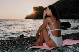 Woman sea yoga. Happy woman meditating in yoga pose on sunset beach, ocean and rock mountains. Motivation and inspirational fit and exercising. Healthy lifestyle outdoors in nature, fitness concept. photo