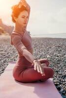 Middle aged well looking woman with black hair, fitness instructor in leggings and tops doing stretching and pilates on yoga mat near the sea. Female fitness yoga routine concept. Healthy lifestyle photo