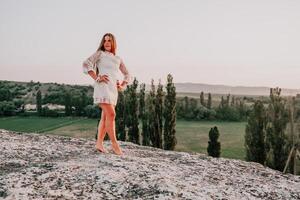 contento mujer en blanco boho vestir en puesta de sol en montañas. romántico mujer con largo pelo en pie con su espalda en el puesta de sol en naturaleza en verano con abierto manos. silueta. naturaleza. puesta de sol. foto