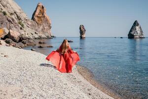 mujer viaje mar. contento turista en rojo vestir disfrutar tomando imagen al aire libre para recuerdos. mujer viajero posando en el rock a mar bahía rodeado por volcánico montañas, compartiendo viaje aventuras viaje foto