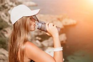 mujer viaje mar. contento turista bebida agua en caliente verano día. mujer viajero mira a el borde de el acantilado en el mar bahía de montañas, compartiendo viaje aventuras viaje foto