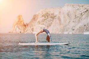 Woman sup yoga. Middle age sporty woman practising yoga pilates on paddle sup surfboard. Female stretching doing workout on sea water. Modern individual female hipster outdoor summer sport activity. photo