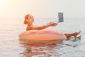 mujer persona de libre dedicación trabajos en ordenador portátil nadando en mar en rosado inflable anillo. contento turista flotante en inflable rosquilla y trabajando en ordenador portátil computadora en calma océano. Lanza libre, remoto trabajando en cualquier sitio foto