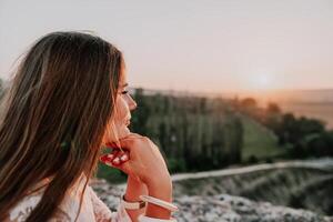 contento mujer en blanco boho vestir en puesta de sol en montañas. romántico mujer con largo pelo en pie con su espalda en el puesta de sol en naturaleza en verano con abierto manos. silueta. naturaleza. puesta de sol. foto