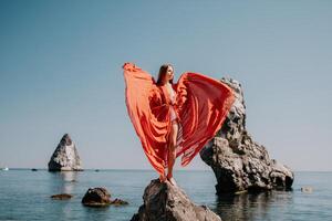 Woman travel sea. Young Happy woman in a long red dress posing on a beach near the sea on background of volcanic rocks, like in Iceland, sharing travel adventure journey photo