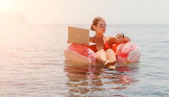 mujer persona de libre dedicación trabajos en ordenador portátil nadando en mar en rosado inflable anillo. contento turista flotante en inflable rosquilla y trabajando en ordenador portátil computadora en calma océano. Lanza libre, remoto trabajando en cualquier sitio foto