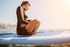 Woman sea laptop. Digital nomad, freelancer with laptop working on sup board at calm sea beach. Happy smiling girl relieves stress from work. Freelance, digital nomad, travel and holidays concept photo