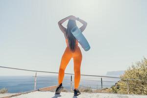 Fitness woman sea. Happy middle aged woman in orange sportswear exercises morning outdoors on yoga mat with laptop in park over ocean beach. Female fitness pilates yoga routine. Healthy lifestyle. photo