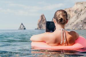 mujer persona de libre dedicación trabajos en ordenador portátil nadando en mar en rosado inflable anillo. contento turista en Gafas de sol flotante en inflable rosquilla y trabajando en ordenador portátil computadora en calma océano. remoto trabajando en cualquier sitio foto