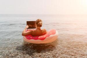 mujer persona de libre dedicación trabajos en ordenador portátil nadando en mar en rosado inflable anillo. contento turista flotante en inflable rosquilla y trabajando en ordenador portátil computadora en calma océano. Lanza libre, remoto trabajando en cualquier sitio foto