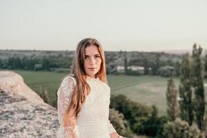 Happy woman in white boho dress on sunset in mountains. Romantic woman with long hair standing with her back on the sunset in nature in summer with open hands. Silhouette. Nature. Sunset. photo