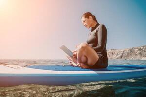 Woman sea laptop. Digital nomad, freelancer with laptop working on sup board at calm sea beach. Happy smiling girl relieves stress from work. Freelance, digital nomad, travel and holidays concept photo