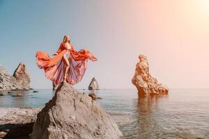 Woman travel sea. Young Happy woman in a long red dress posing on a beach near the sea on background of volcanic rocks, like in Iceland, sharing travel adventure journey photo