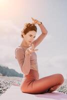 Middle aged well looking woman with black hair, fitness instructor in leggings and tops doing stretching and pilates on yoga mat near the sea. Female fitness yoga routine concept. Healthy lifestyle photo