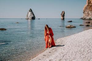 Woman travel sea. Happy tourist in red dress enjoy taking picture outdoors for memories. Woman traveler posing on the rock at sea bay surrounded by volcanic mountains, sharing travel adventure journey photo