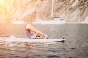 Woman sup yoga. Middle age sporty woman practising yoga pilates on paddle sup surfboard. Female stretching doing workout on sea water. Modern individual female hipster outdoor summer sport activity. photo