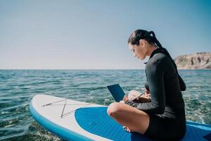 Woman sea laptop. Digital nomad, freelancer with laptop working on sup board at calm sea beach. Happy smiling girl relieves stress from work. Freelance, digital nomad, travel and holidays concept photo