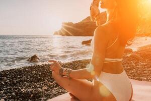 Woman sea yoga. Happy woman in white swimsuit and boho style braclets practicing outdoors on yoga mat by sea on sunset. Women yoga fitness routine. Healthy lifestyle, harmony and meditation photo