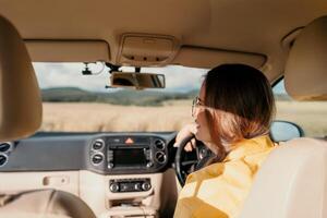 un mujer en un amarillo camisa es conducción un coche. ella es vistiendo lentes y tiene su manos en el direccion rueda foto