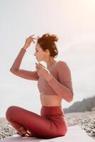 Middle aged well looking woman with black hair, fitness instructor in leggings and tops doing stretching and pilates on yoga mat near the sea. Female fitness yoga routine concept. Healthy lifestyle photo