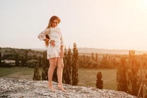 Happy woman in white boho dress on sunset in mountains. Romantic woman with long hair standing with her back on the sunset in nature in summer with open hands. Silhouette. Nature. Sunset. photo