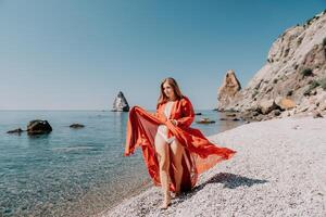 Woman travel sea. Happy tourist in red dress enjoy taking picture outdoors for memories. Woman traveler posing on the rock at sea bay surrounded by volcanic mountains, sharing travel adventure journey photo