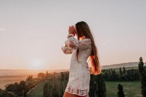 Happy woman in white boho dress on sunset in mountains. Romantic woman with long hair standing with her back on the sunset in nature in summer with open hands. Silhouette. Nature. Sunset. photo
