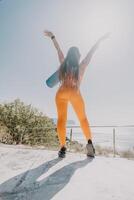 Fitness woman sea. Happy middle aged woman in orange sportswear exercises morning outdoors on yoga mat with laptop in park over ocean beach. Female fitness pilates yoga routine. Healthy lifestyle. photo