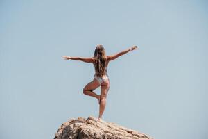 Woman sea yoga. Back view of free calm bliss satisfied woman with long hair standing on top rock with yoga position against of sky by the sea. Healthy lifestyle outdoors in nature, fitness concept. photo