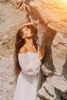 mujer verano viaje mar. contento dama con largo pelo en blanco vestir disfrutar tomando foto al aire libre para recuerdos. mujer viajero posando en playa a mar rodeado por volcánico montañas, compartiendo emociones