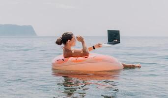 Woman freelancer works on laptop swimming in sea on pink inflatable ring. Happy tourist floating on inflatable donut and working on laptop computer in calm ocean. Freelance, remote working anywhere photo