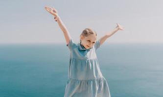 A young girl is standing on a beach, wearing a blue dress and holding her arms up in the air. Concept of joy and freedom, as the girl appears to be enjoying her time by the ocean. photo