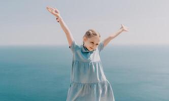 un joven niña es en pie en un playa, vistiendo un azul vestir y participación su brazos arriba en el aire. concepto de alegría y libertad, como el niña aparece a ser disfrutando su hora por el océano. foto