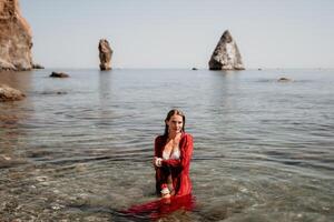 Woman travel sea. Happy tourist in red dress enjoy taking picture outdoors for memories. Woman traveler posing in sea beach, surrounded by volcanic mountains, sharing travel adventure journey photo