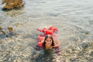 mujer viaje mar. contento turista en rojo vestir disfrutar tomando imagen al aire libre para recuerdos. mujer viajero posando en mar playa, rodeado por volcánico montañas, compartiendo viaje aventuras viaje foto