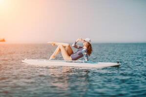 Woman sup yoga. Middle age sporty woman practising yoga pilates on paddle sup surfboard. Female stretching doing workout on sea water. Modern individual female hipster outdoor summer sport activity. photo