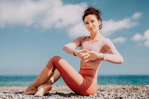 Middle aged well looking woman with black hair, fitness instructor in leggings and tops doing stretching and pilates on yoga mat near the sea. Female fitness yoga routine concept. Healthy lifestyle photo