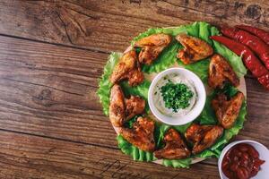 delicious grilled chicken wings with garlic and tomato sauce with lettuce on a round board on wooden rustic background top view, witi copyspace photo