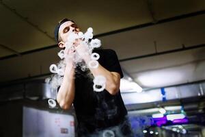 Man in cap smoke an electronic cigarette and releases clouds of vapor performing various kind of vaping tricks photo