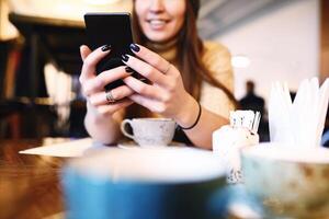 mujer mecanografía texto mensaje en inteligente teléfono en un cafetería. recortado imagen de joven mujer sentado a un mesa con un café utilizando móvil teléfono. tonificado selectivo enfocar. foto