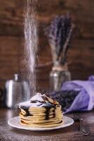 Stack of delicious pancakes with chocolate, honey, slices of banana and castor sugar on plate on wooden background, cinnamon sticks and bunch of lavender. photo