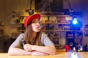 Young pretty woman in red cap smoke an electronic cigarette at the vape shop photo