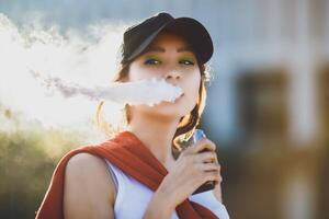 Pretty young hipster asian woman in black hat vape ecig, vaping device at the sunset. Toned image. Closeup. photo