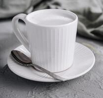 blanco taza de espumoso Leche café en un texturizado mesa con un cuchara y verde hojas foto