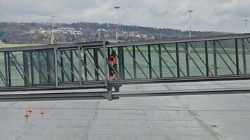 arbeider Bij luchthaven terminal loopbrug, arbeider in hoog zichtbaarheid oranje uniform wandelen langs een luchthaven terminal glas loopbrug, reflecterend een bezig vliegveld en weelderig groen landschap in de achtergrond. video