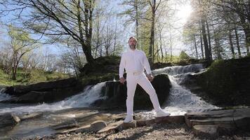 un hombre en blanco ropa canta en el bosque en contra el antecedentes de un cascada y naturaleza video