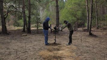 On an autumn morning, builders drill the soil in a forest clearing to install a concrete foundation. The ground is marked with several small stakes, indicating a methodical approach to their work. video